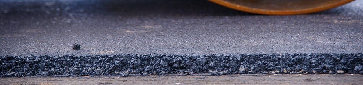 A freshly laid asphalt road by Palm Beach Asphalt is in the process of being compacted by a roller. The image shows a close-up view of the smooth top layer and the rough, lower layer, capturing the precision of asphalt paving in stunning detail.