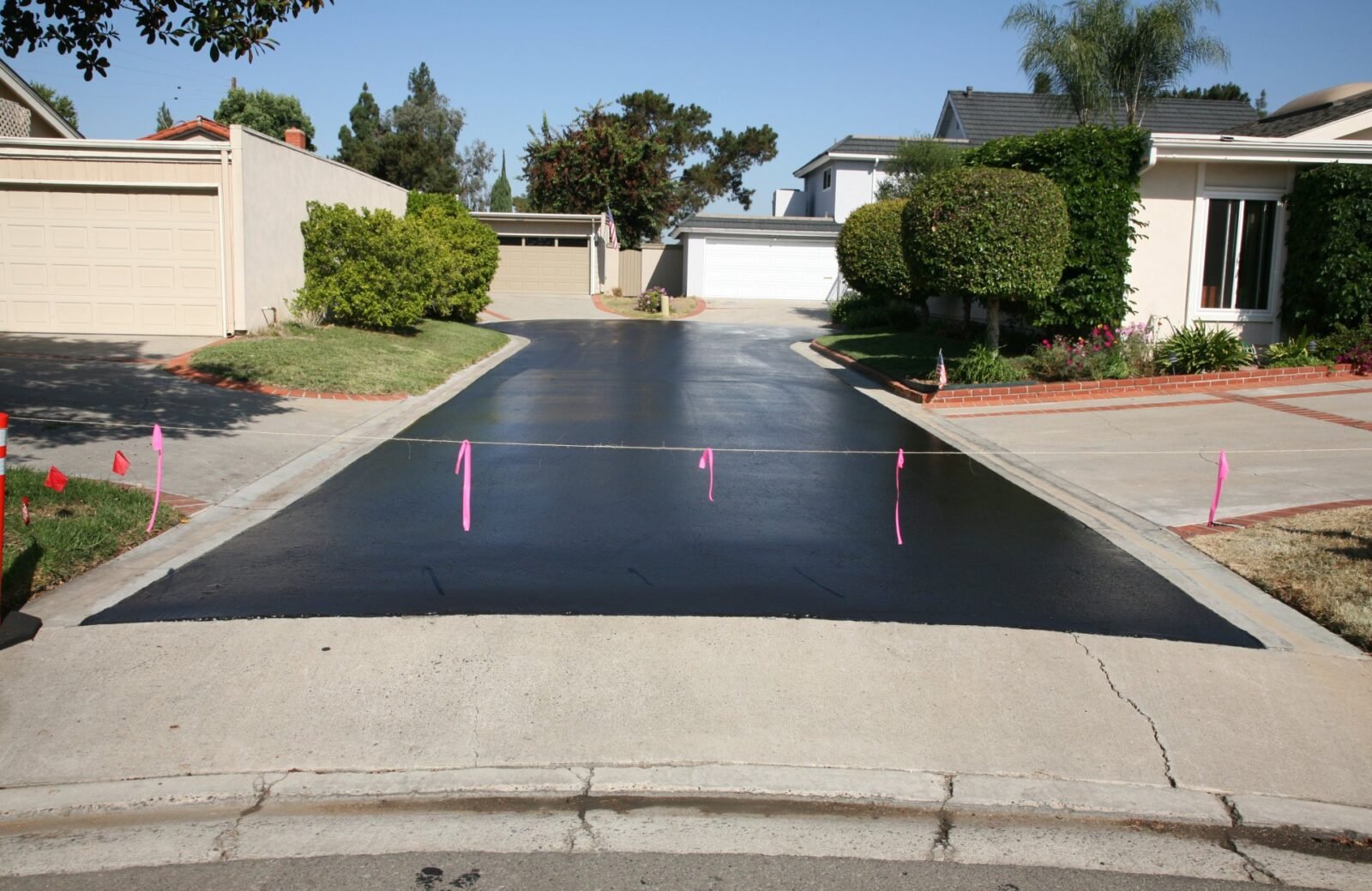A freshly paved black asphalt driveway by Palm Beach Asphalt is roped off with pink ribbons. It is situated between two white houses with well-kept lawns and shrubs. There is a garage at the end of the driveway. The sky is clear and blue.