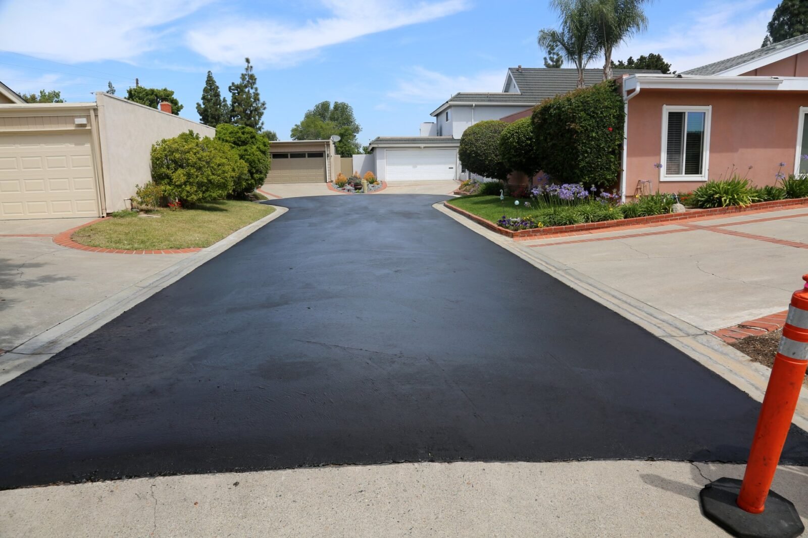 A freshly paved asphalt driveway, expertly completed by Palm Beach Asphalt, leading to two white garages and a beige garage in a residential neighborhood. The driveway is bordered by curbs and landscaped with grass, flowers, and a palm tree. An orange traffic cone is visible in the foreground. Get your free quote today!