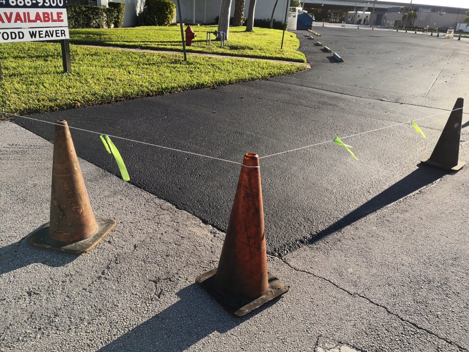 Two traffic cones are connected by a thin rope with yellow flags marking off a section of Palm Beach Asphalts freshly paved area next to a grassy patch. In the background, a sign reads AVAILABLE, hinting at the contractor's meticulous asphalt paving work.