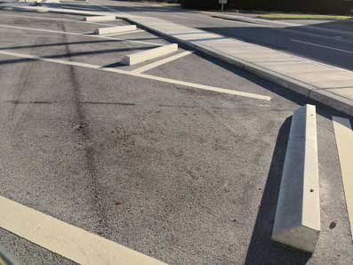 A nearly empty parking lot with marked parking spaces and concrete wheel stops. The asphalt paving shows faint tire marks, hinting at recent use. Bright sunlight casts shadows from the surrounding elements onto the pavement, bringing a touch of Delray Beach to this serene scene.