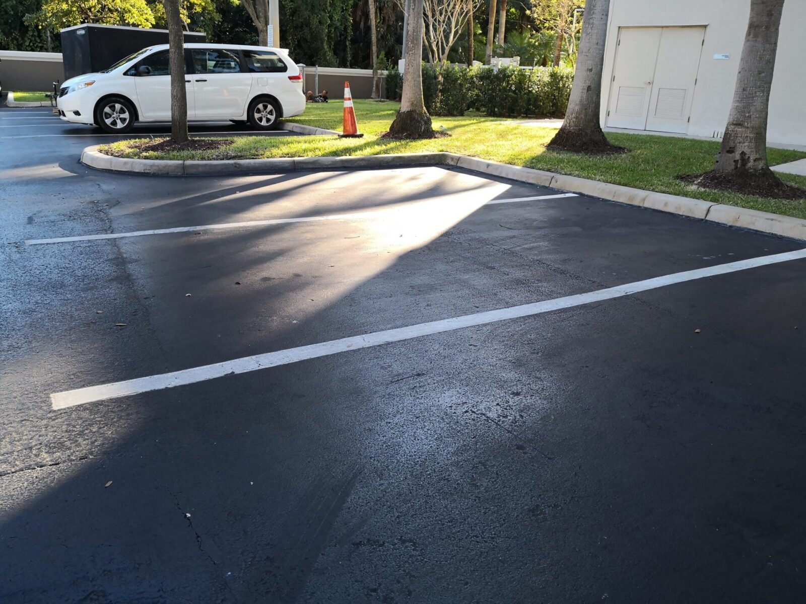 A small parking lot with a few empty spaces, ideal for fresh asphalt paving by Palm Beach Asphalt. A white minivan is parked next to the curb with an orange traffic cone placed in front of it. Trees and a small building entrance are visible in the background. Get your free quote today!