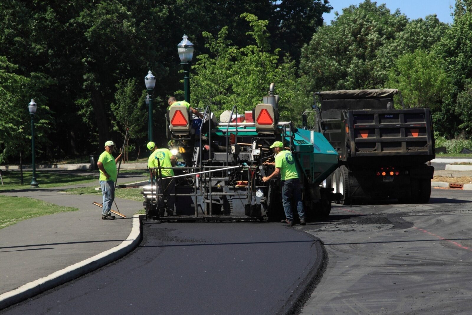 A crew of workers in bright green shirts and helmets is operating machinery for asphalt paving on a road in Riviera Beach. A large dump truck from Palm Beach Asphalt is unloading asphalt as workers use tools to smooth and pave the surface. Trees and street lamps are visible in the background.