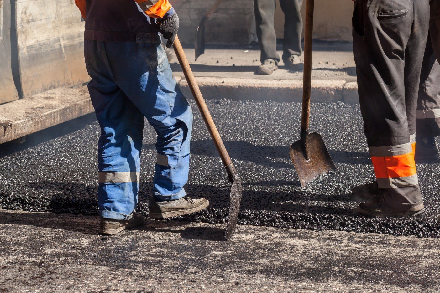 Three road workers from Palm Beach Asphalt are engaged in asphalt paving. They wear safety gear including heavy boots and gloves. Two hold shovels, while the third uses a rake to spread the fresh black asphalt. They stand on top of the new layer being applied to the road surface by their contractor.