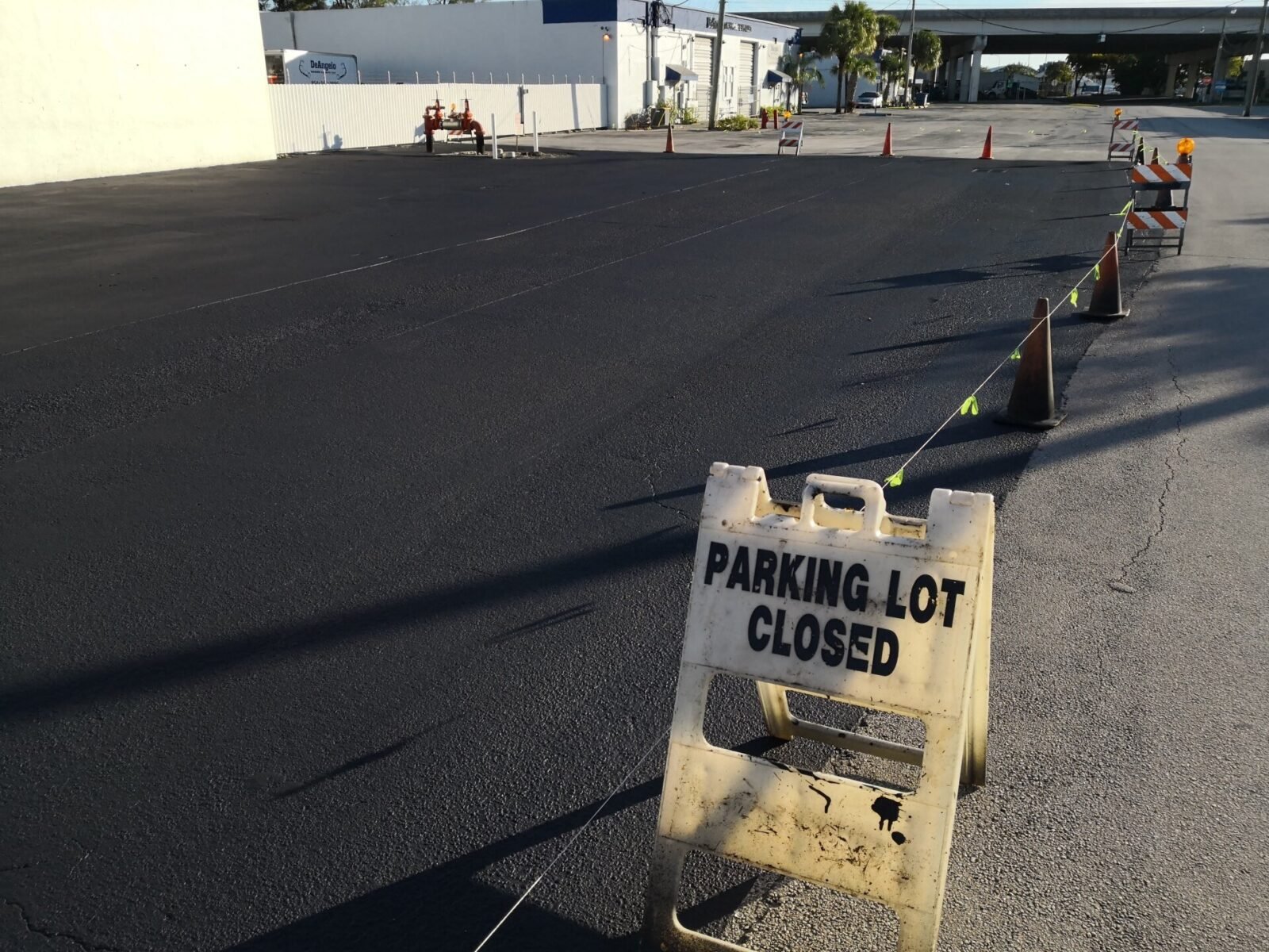 Parking Lot Closed in the foreground of an image showing an empty, freshly paved parking lot by Palm Beach Asphalt. Traffic cones and a barricade are visible, blocking off the area. There's a building and some trees in the background under a clear Boynton Beach FL sky.