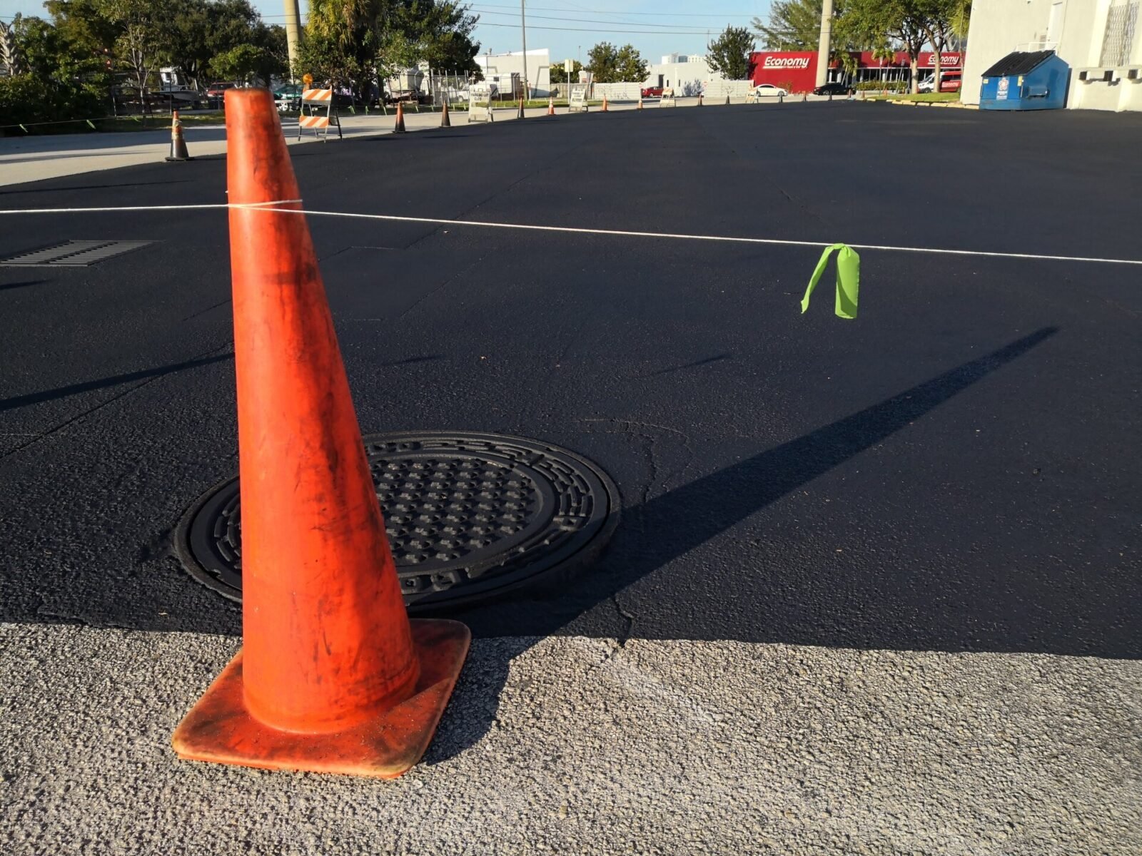 An orange traffic cone is positioned on a freshly paved, black asphalt surface by Palm Beach Asphalt next to a manhole cover. A white string with a small, green ribbon is attached to the cone, extending to the right. Trees, a few buildings, and traffic cones are visible in the distance.