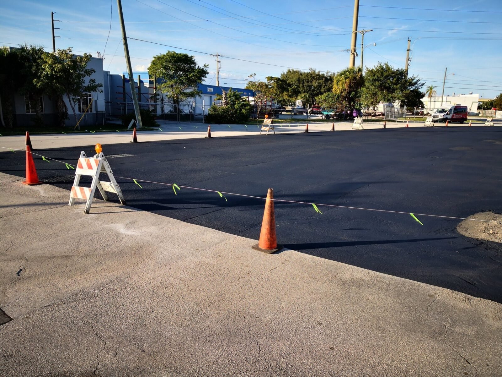 A newly paved black asphalt area is cordoned off with traffic cones and caution tape. There is a white barricade with orange stripes on the left, indicating recent asphalt paving by Palm Beach Asphalt. Vehicles and buildings are visible in the background on a sunny Riviera Beach day.