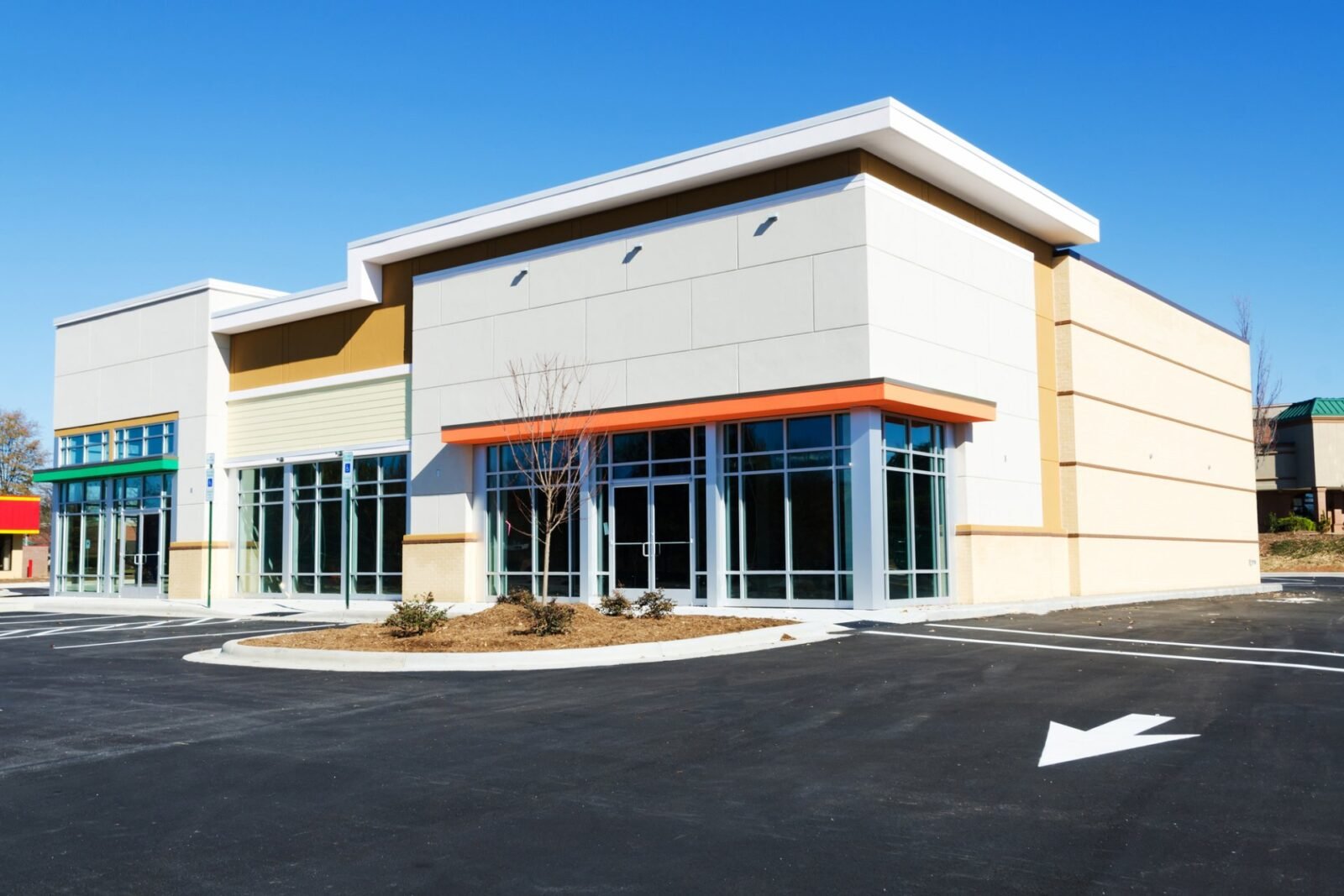 A new, modern commercial building with large windows and a flat roof constructed by a skilled contractor. The storefront features a mix of beige, white, and orange accents. The surrounding area has a spacious, freshly paved parking lot courtesy of Palm Beach Asphalt, with a few small shrubs and a clear blue sky above.