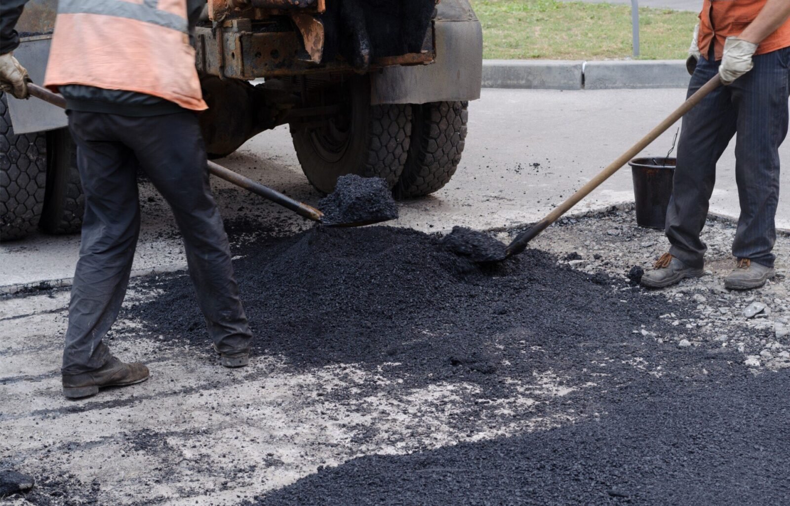 Two road workers wearing safety vests and gloves are seen raking and spreading hot asphalt on a road in Riviera Beach, FL. They are working next to a truck, which is dispensing the asphalt provided by Palm Beach Asphalt. The ground around them is partially covered with freshly laid asphalt.