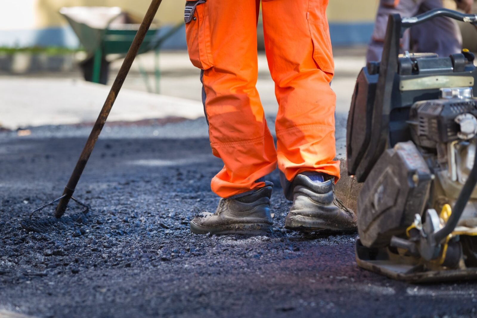 A construction worker wearing bright orange pants and sturdy boots stands on freshly laid asphalt while holding a tool. Another piece of construction equipment is visible on the right side of the image, alongside a wheelbarrow in the blurred background, showcasing Palm Beach Asphalt's meticulous craftsmanship.