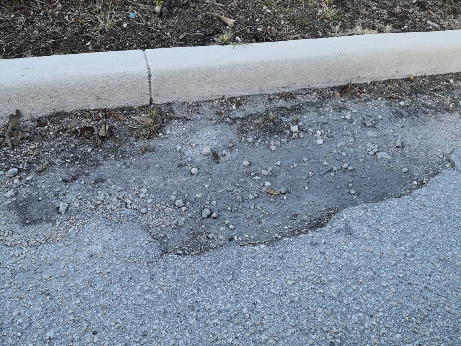 A pothole with broken asphalt and loose gravel is situated near a curb in an asphalt-paved area. The surrounding surface shows signs of wear and deterioration, with small patches of grass and dirt visible along the edge of the curb—a reminder that quality asphalt paving services like those provided by Palm Beach Asphalt are essential.